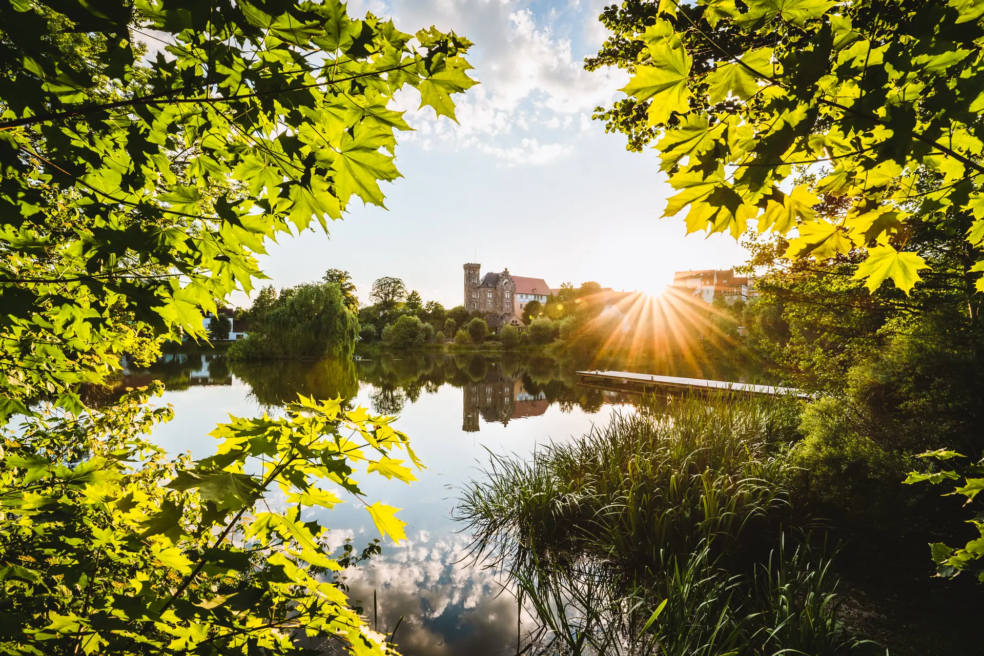 Schloss Ronneburg hinter einem Teich im Gegenlicht der Abendsonne, gerahmt von Ahornblättern